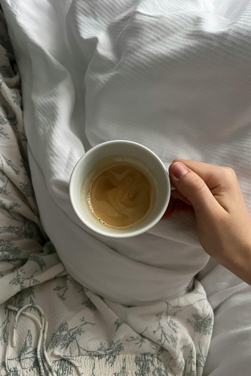 A hand holding a small cup of coffee while resting in bed under soft white linen.