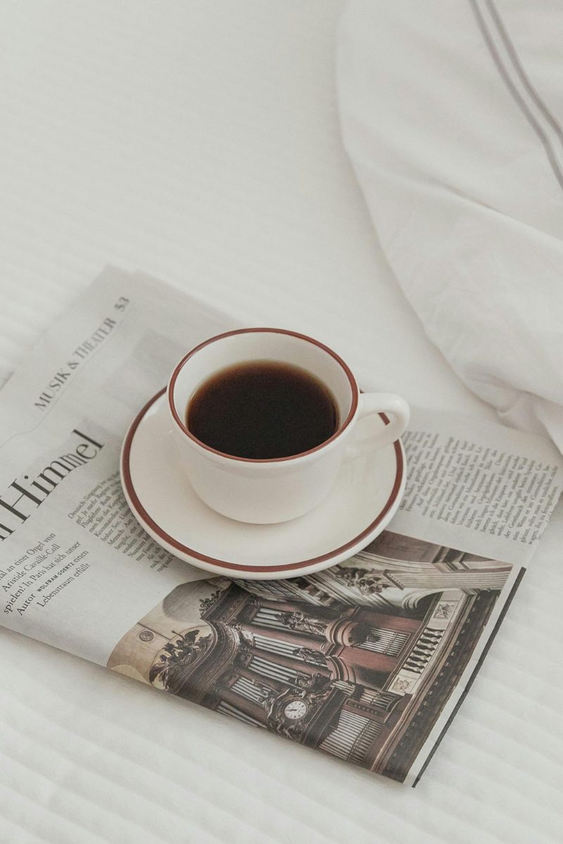 A cup of coffee on a saucer resting on an open magazine, on white bedlinen.
