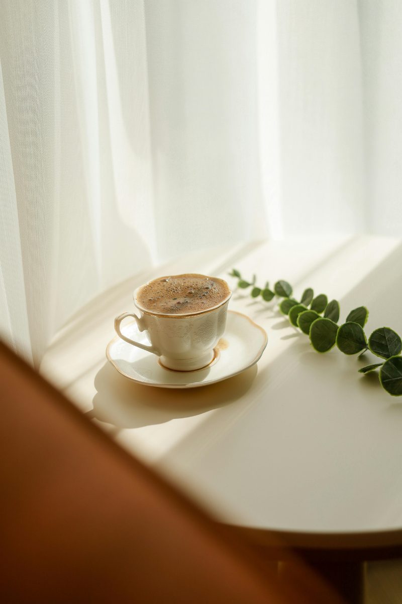 A delicate cup and saucer on a windowsill beside a sprig of eucalyptus, sheer curtains diffusing the light.