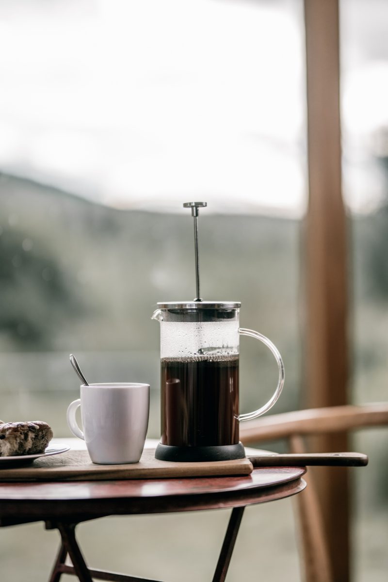 A glass coffee press and a white mug on a small table by a window, hills softly blurred beyond.