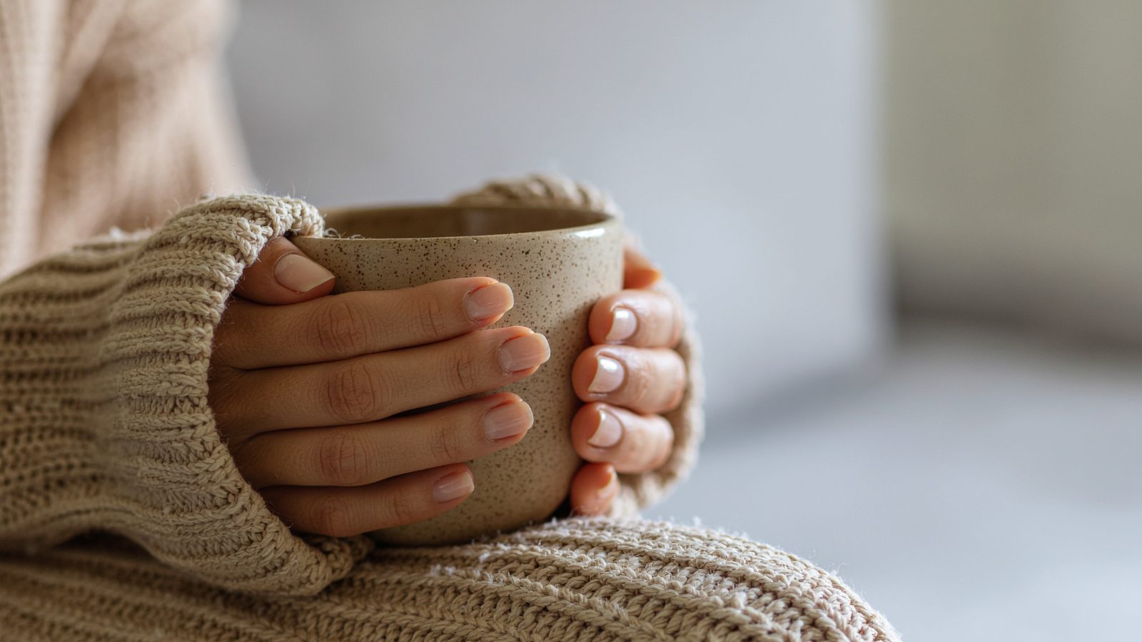 Two hands in a knitted jumper wrapped around a warm speckled mug, soft window light behind.