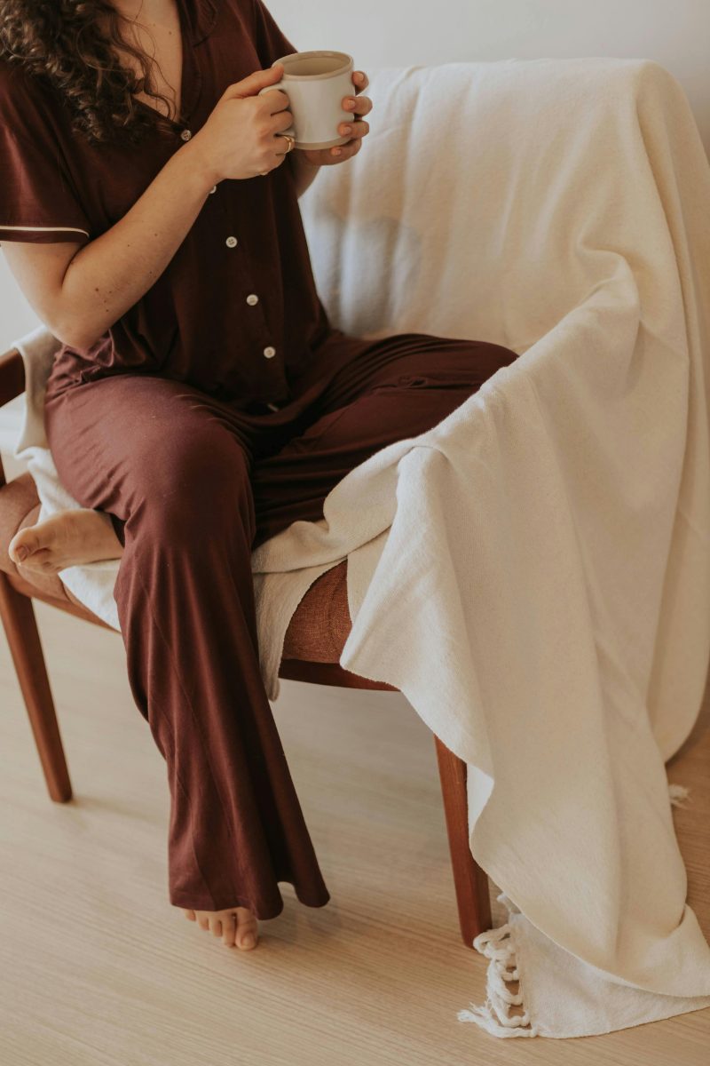 A woman in soft loungewear sits with a warm mug and a cream throw over her lap.