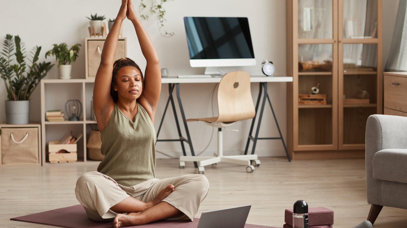 A woman sits cross-legged on a mat in a calm living room, arms stretched overhead, eyes closed, in soft morning light.